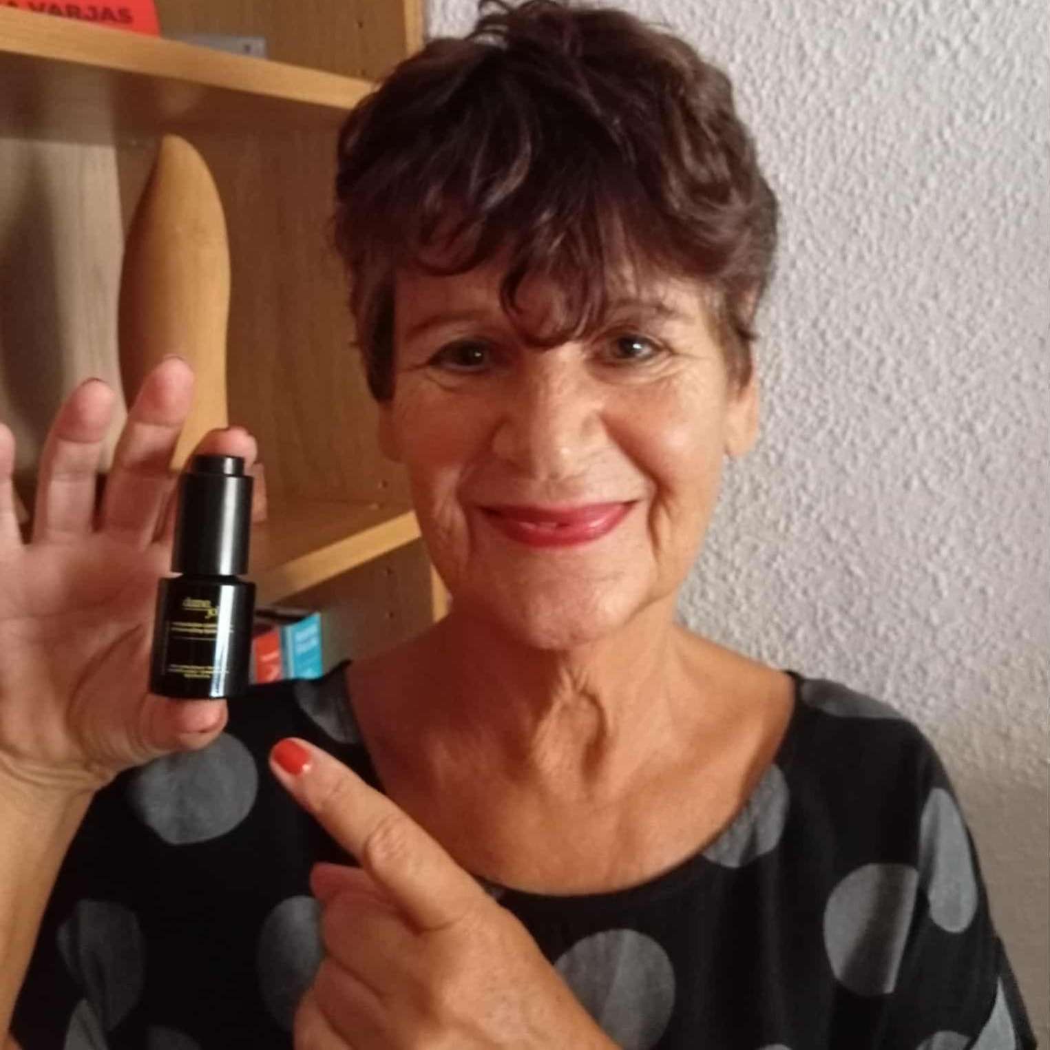 A woman smiling and pointing at the Dame Jo! Executive Radiance Antipollution Lipid Serum, holding the sleek black bottle in a home setting to showcase her skincare routine.