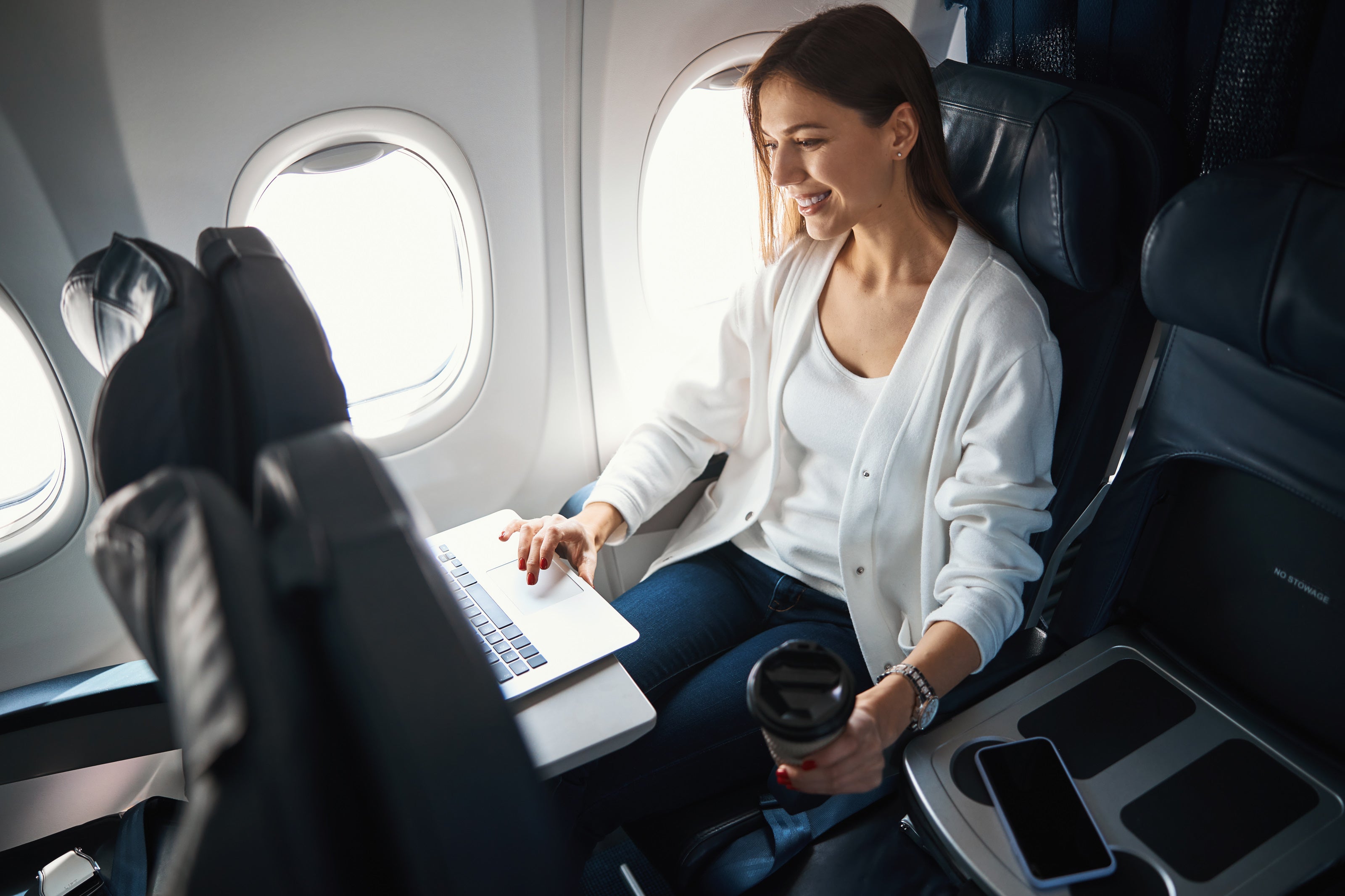 Woman using a laptop on an airplane with a cup and phone in hand.