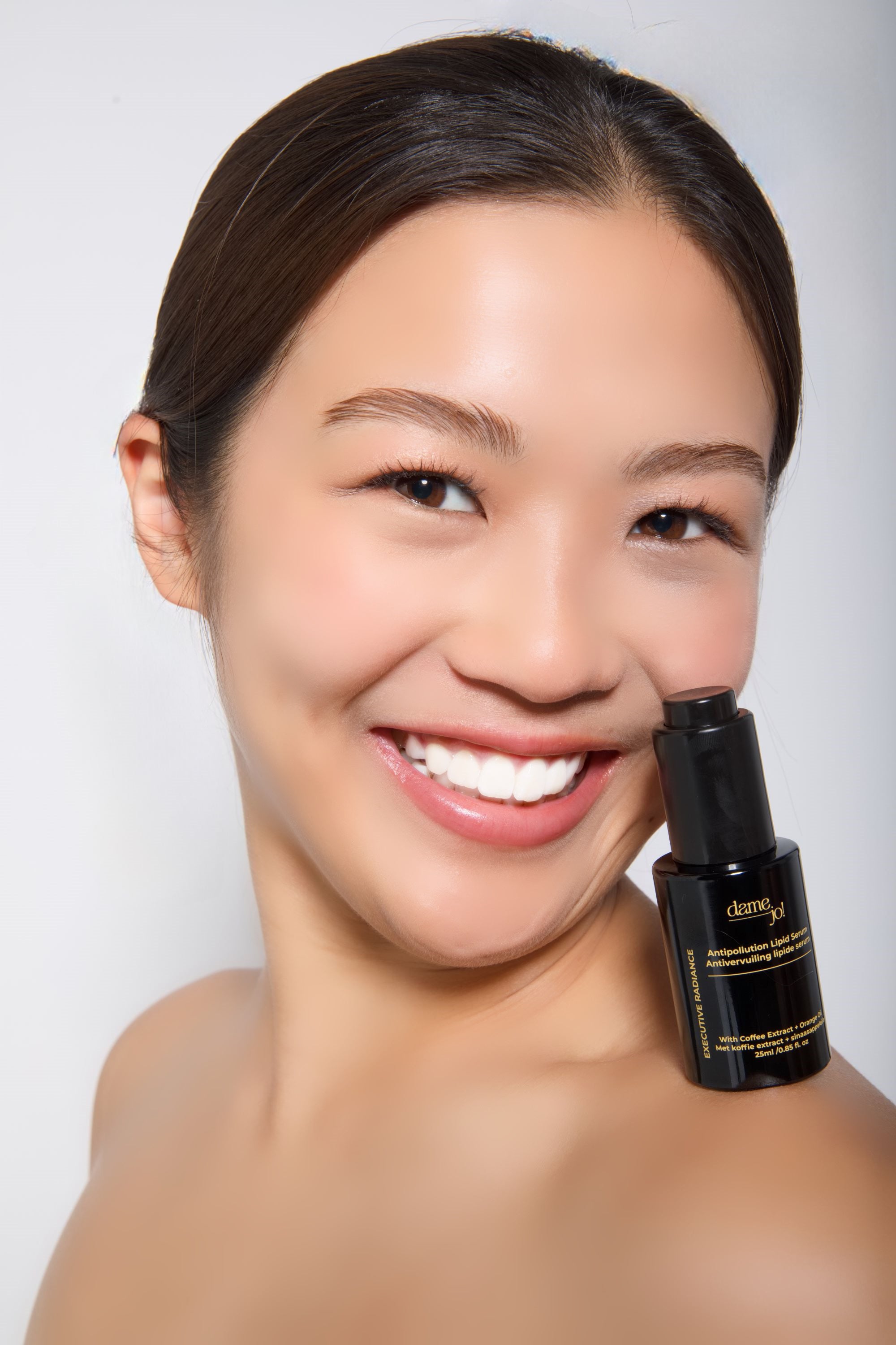 Model smiling with Dame Jo! serum applied to shoulder, radiating confidence and glow.