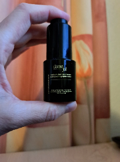 Close-up of a hand holding the Dame Jo! Executive Radiance Antipollution Lipid Serum; elegant black packaging with coffee extract and orange oil formula shown against a warm indoor backdrop.