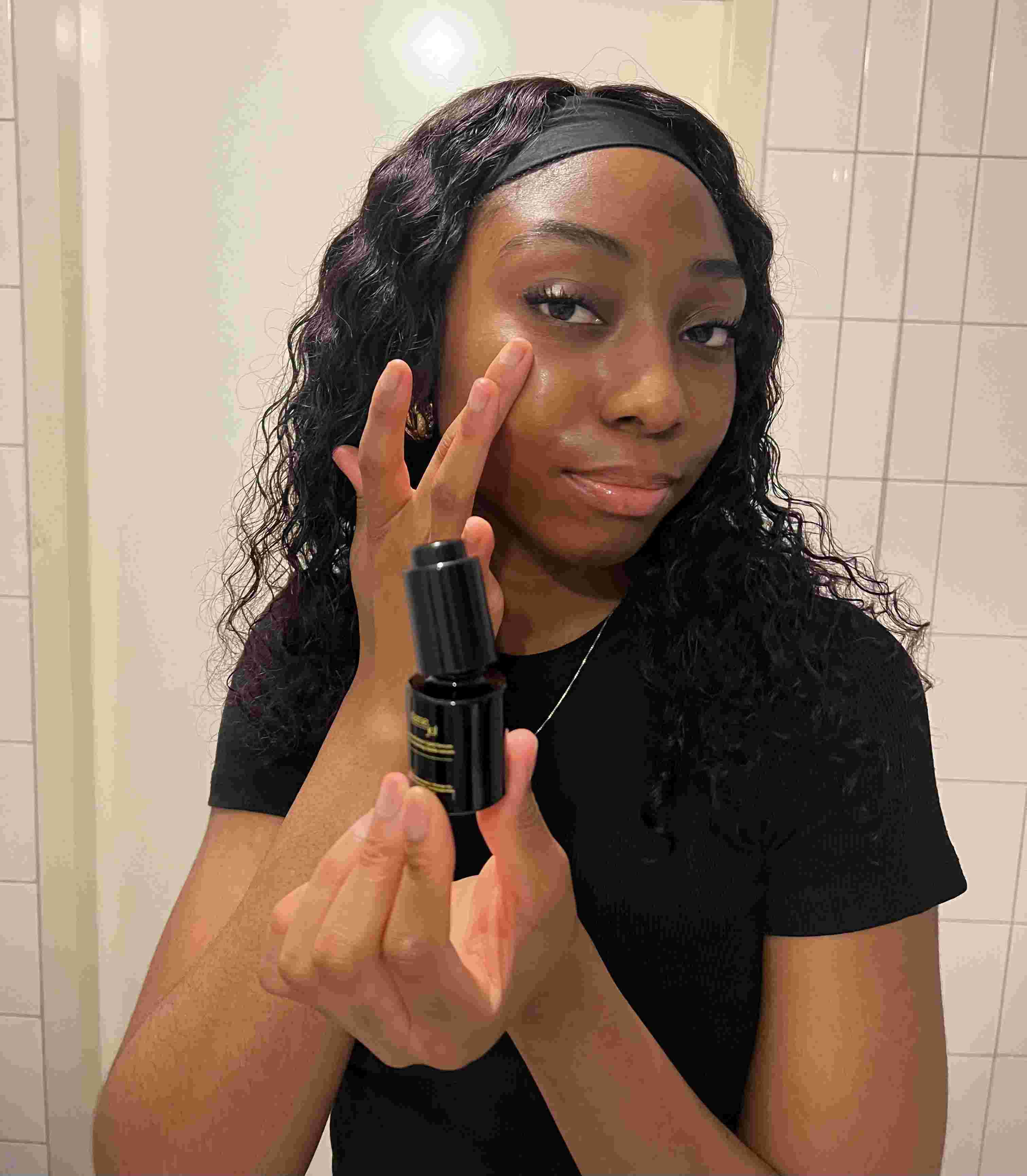 A woman applying Dame Jo! Executive Radiance Antipollution Lipid Serum to her cheek, holding the sleek black bottle in a bathroom setting; demonstrating a natural skincare routine for radiant, pollution-protected skin.